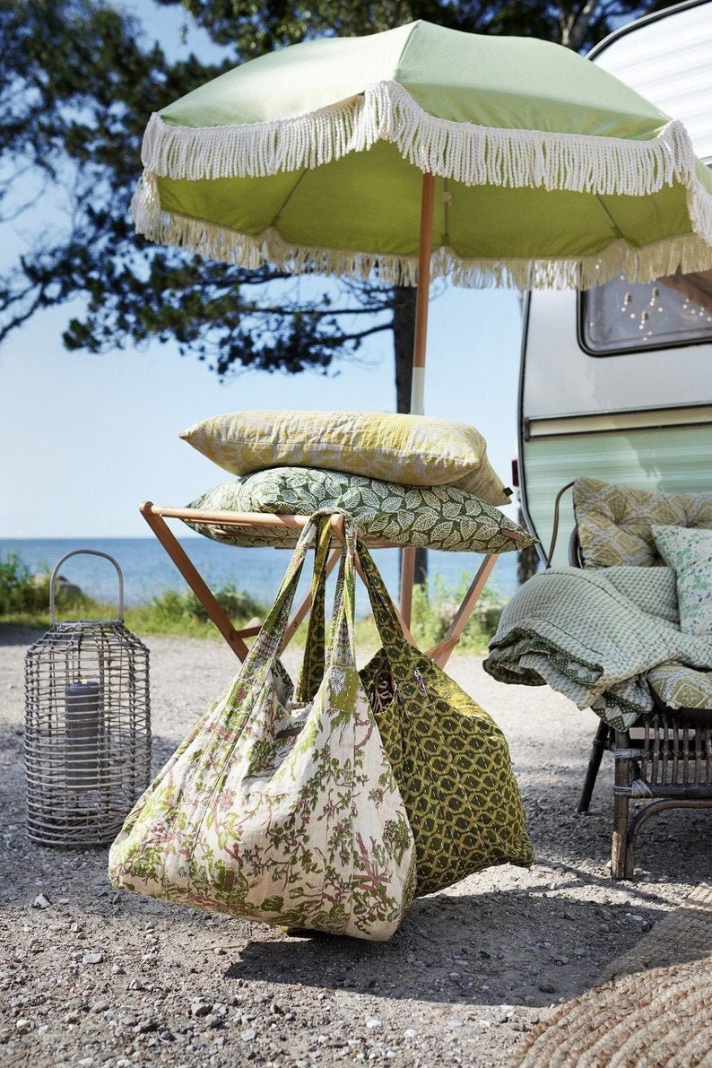 May include: A green and white striped beach umbrella with fringe trim, two green and white patterned tote bags, and a variety of green and white patterned pillows and blankets. The scene is set up in front of a white camper van.