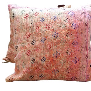 Peut inclure: Un coussin carré décoratif avec un motif tissé texturé. Le coussin présente une palette de couleurs pêche et crème avec un motif de losanges rouges, bleus et noirs. Des accents de cuir sont visibles.
