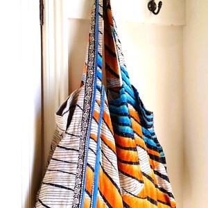 May include: A large, quilted tote bag with a white background and orange, blue, and black stripes. The bag has a blue and silver trim and is hanging on a hook.