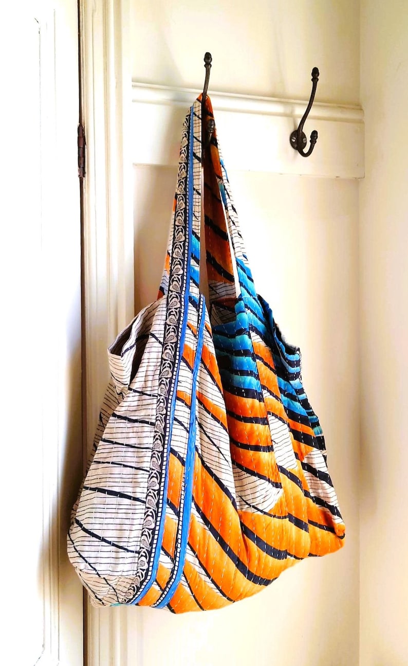 May include: A large, quilted tote bag with a white background and orange, blue, and black stripes. The bag has a blue and silver trim and is hanging on a hook.