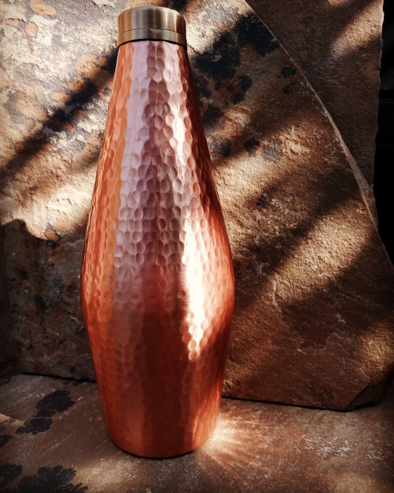 May include: A hammered copper water bottle with a wide mouth and a copper lid. The bottle is sitting on a brown stone surface.