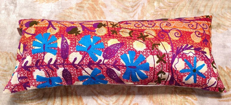 May include: A long, rectangular pillow with a colorful floral pattern. The pillow is made of a soft, textured fabric and features a variety of blue, purple, and orange flowers on a red background.
