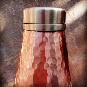 May include: A hammered copper water bottle with a silver lid. The bottle has a textured, hammered finish and a rounded shape.