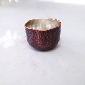 Puede incluir: Una pequeña taza de cobre martillada con un interior plateado. La taza tiene una forma redondeada y una superficie texturizada.