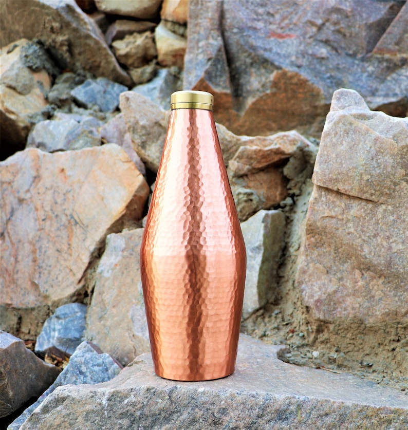 May include: A hammered copper water bottle with a brass lid. The bottle is standing on a rock.