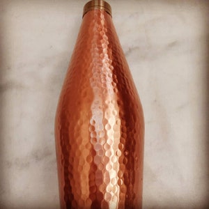 May include: A hammered copper water bottle with a brass lid. The bottle has a rounded, slightly tapered shape.