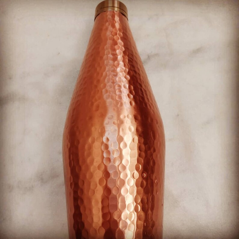 May include: A hammered copper water bottle with a brass lid. The bottle has a rounded, slightly tapered shape.