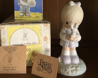 May include: A collection of Precious Moments figurines and boxes. One figurine depicts a child holding a bunny. Another figurine is an astronaut. Boxes and tags with text: "Wishing You A Happy Easter" and "The Club That's Out Of This World."