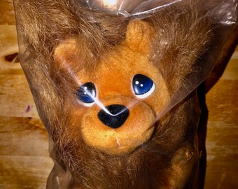 May include: A brown plush lion with blue eyes and a black nose. The lion is packaged in a clear plastic bag with a warning label that reads "WARNING: To avoid danger of suffocation, keep away from babies and children. Do not use in cribs, carriages or play pens."