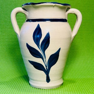 May include: Cream-colored ceramic vase with two handles and a dark blue leaf design. The vase has a ruffled rim and a dark blue band around the top. The vase is on a green background.