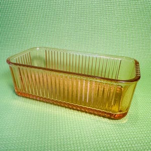 May include: A rectangular, amber-colored glass loaf pan with vertical ribbed detailing. The vintage bakeware sits on a green surface. The pan's design features a slightly textured rim and a classic, functional shape.