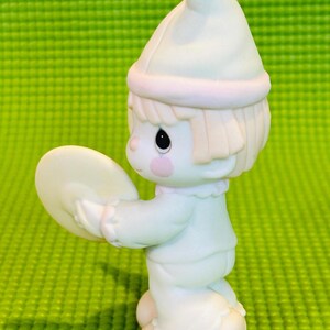 May include: A small porcelain figurine of a clown holding a cymbal. The clown is wearing a white hat with a pink band and a white and blue outfit. The figurine is standing on a green surface.
