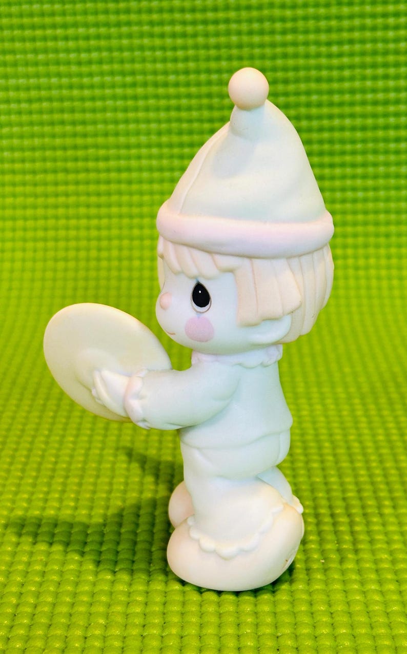 May include: A small porcelain figurine of a clown holding a cymbal. The clown is wearing a white hat with a pink band and a white and blue outfit. The figurine is standing on a green surface.