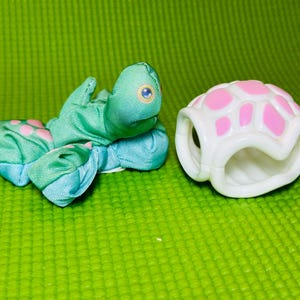 May include: A small green fabric turtle toy with pink spots and blue eyes, positioned next to a separate white plastic turtle shell with pink accents. The fabric turtle has a soft texture, while the shell is a hard, hollow, molded piece.