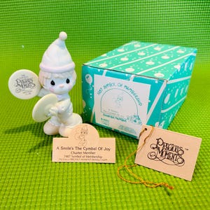 May include: A white porcelain figurine of a smiling angel playing a cymbal. The angel is wearing a white hat and a blue and white outfit. The figurine is part of a Precious Moments collection and is labeled "A Smile's The Cymbal Of Joy, Charter Member, 1987 Symbol of Membership".