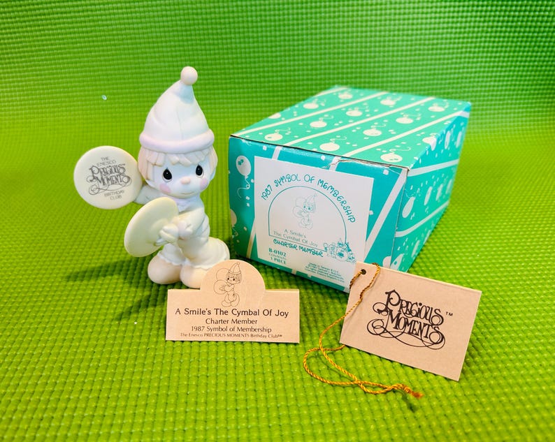 May include: A white porcelain figurine of a smiling angel playing a cymbal. The angel is wearing a white hat and a blue and white outfit. The figurine is part of a Precious Moments collection and is labeled "A Smile's The Cymbal Of Joy, Charter Member, 1987 Symbol of Membership".