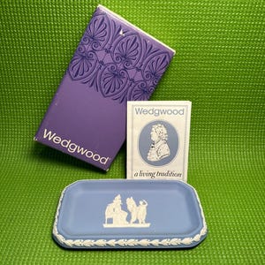 May include: A blue rectangular Wedgwood trinket dish with a white relief design, a purple box with a decorative pattern, and a small card with a portrait and the words "a living tradition". The background is green.