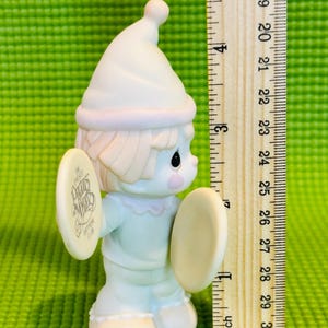 May include: A white porcelain figurine of a clown holding cymbals. The clown is wearing a blue and white outfit and a white hat. The cymbal has the text "The Precious Moments Birthday Club" on it.