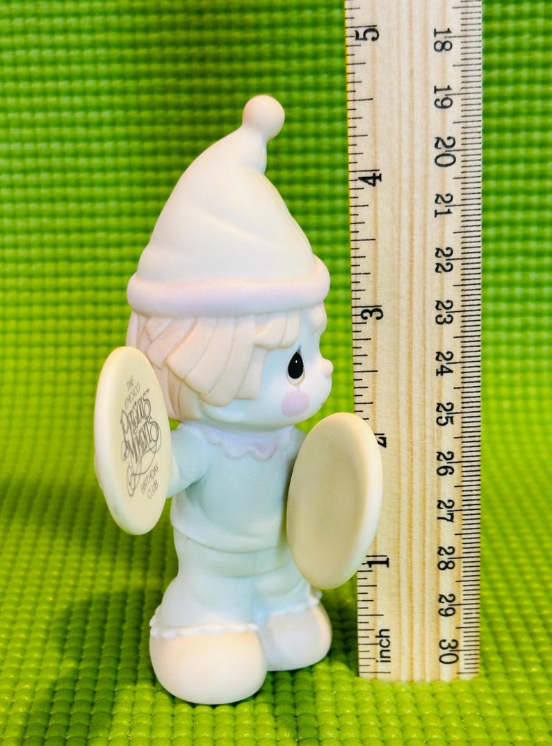 May include: A white porcelain figurine of a clown holding cymbals. The clown is wearing a blue and white outfit and a white hat. The cymbal has the text "The Precious Moments Birthday Club" on it.