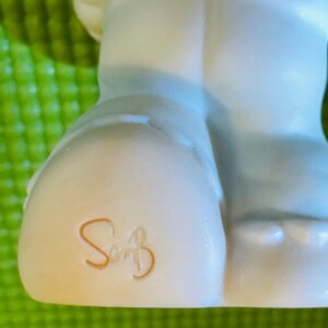 May include: A white ceramic figurine of a person wearing blue pants. The figurine has a signature on the bottom that reads "So" with a flourish.