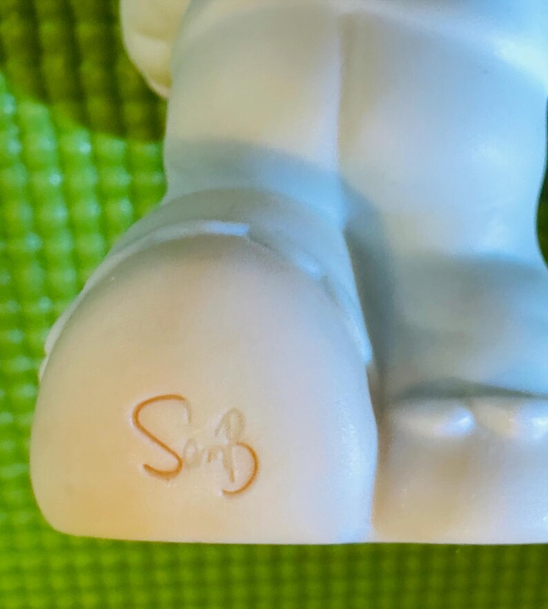 May include: A white ceramic figurine of a person wearing blue pants. The figurine has a signature on the bottom that reads "So" with a flourish.