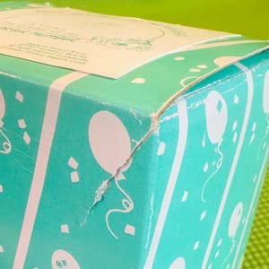 May include: A turquoise blue gift box with a white label that reads "A Classic Series by The Country Rose Collection". The box is decorated with white balloons and confetti.