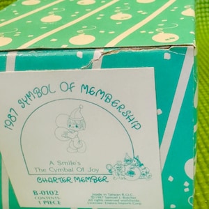 May include: A white box with a green and white pattern on the top. The box has a sticker on the front with a cartoon angel holding balloons. The sticker says "1987 Symbol of Membership, A Smile's The Cymbal Of Joy, Charter Member, B-0102 Contents 1 Piece".