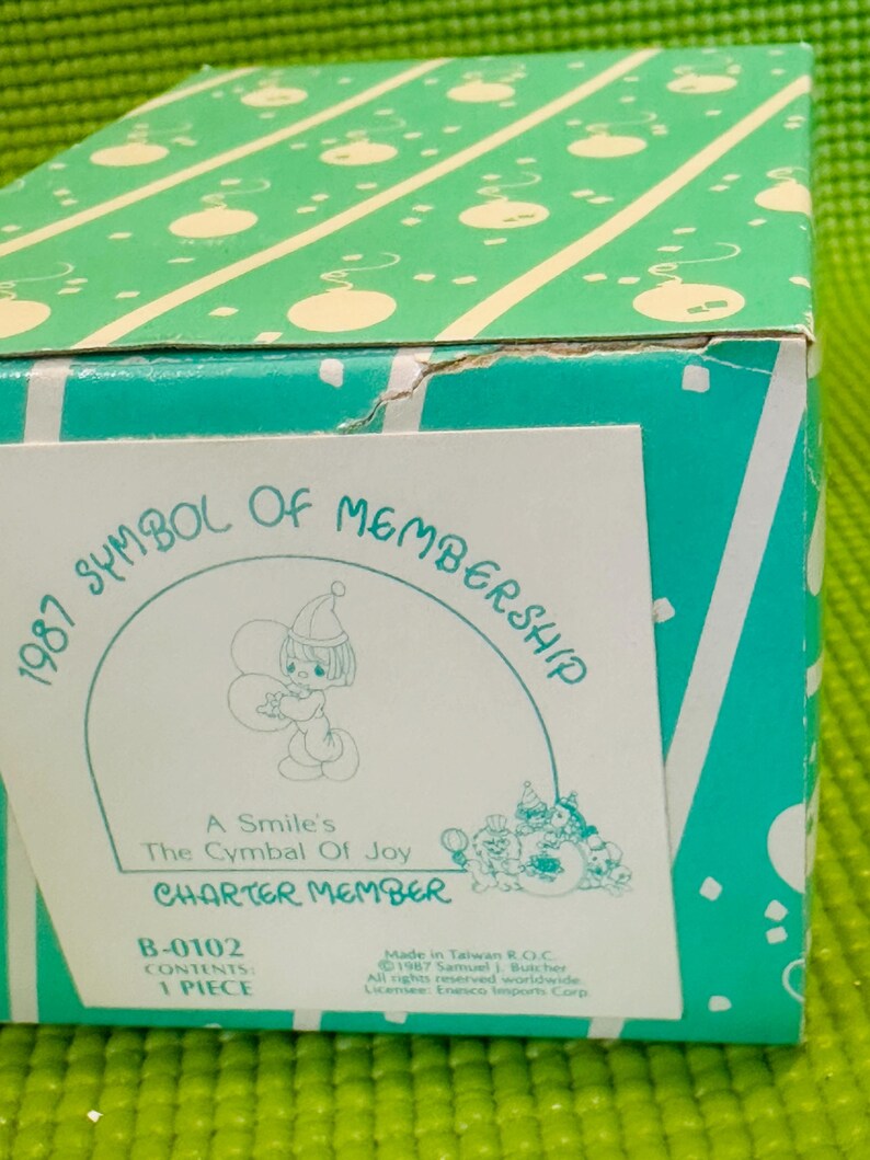 May include: A white box with a green and white pattern on the top. The box has a sticker on the front with a cartoon angel holding balloons. The sticker says "1987 Symbol of Membership, A Smile's The Cymbal Of Joy, Charter Member, B-0102 Contents 1 Piece".