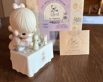 May include: A porcelain figurine of a young girl holding a small doll. The girl is sitting at a table with a small cake on it. The figurine is part of the Precious Moments collection and is titled "My Happiness".