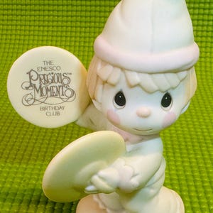 May include: A white porcelain figurine of a boy wearing a white hat and holding a cymbal. The figurine is part of the Precious Moments Birthday Club collection.