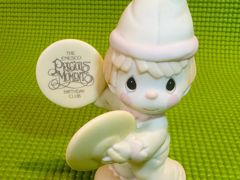 May include: A white porcelain figurine of a boy wearing a white hat and holding a cymbal. The figurine is part of the Precious Moments Birthday Club collection.