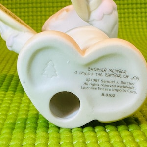 May include: White porcelain figurine of an angel playing a cymbal. The figurine is a charter member of the "A Smile's the Symbol of Joy" collection. The base of the figurine has the text "Charter Member A Smile's the Symbol of Joy &copy; 1987 Samuel J. Butcher All rights reserved worldwide. Licensee Enesco Imports Corp. B-0102"