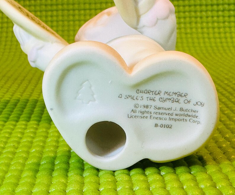 May include: White porcelain figurine of an angel playing a cymbal. The figurine is a charter member of the "A Smile's the Symbol of Joy" collection. The base of the figurine has the text "Charter Member A Smile's the Symbol of Joy &copy; 1987 Samuel J. Butcher All rights reserved worldwide. Licensee Enesco Imports Corp. B-0102"