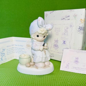 May include: A Precious Moments figurine of a girl in a bonnet, seated and holding a needle. The figurine is on a white base, with a small pot and stones. The box and cards are also visible, with the text "Always Take Time To Pray".