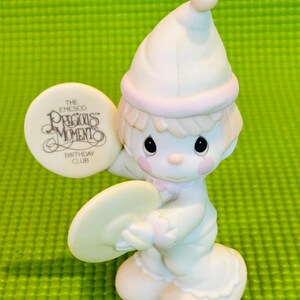 May include: A white porcelain figurine of a boy wearing a white and pink hat, holding a white cymbal. The figurine is part of the Precious Moments Birthday Club.