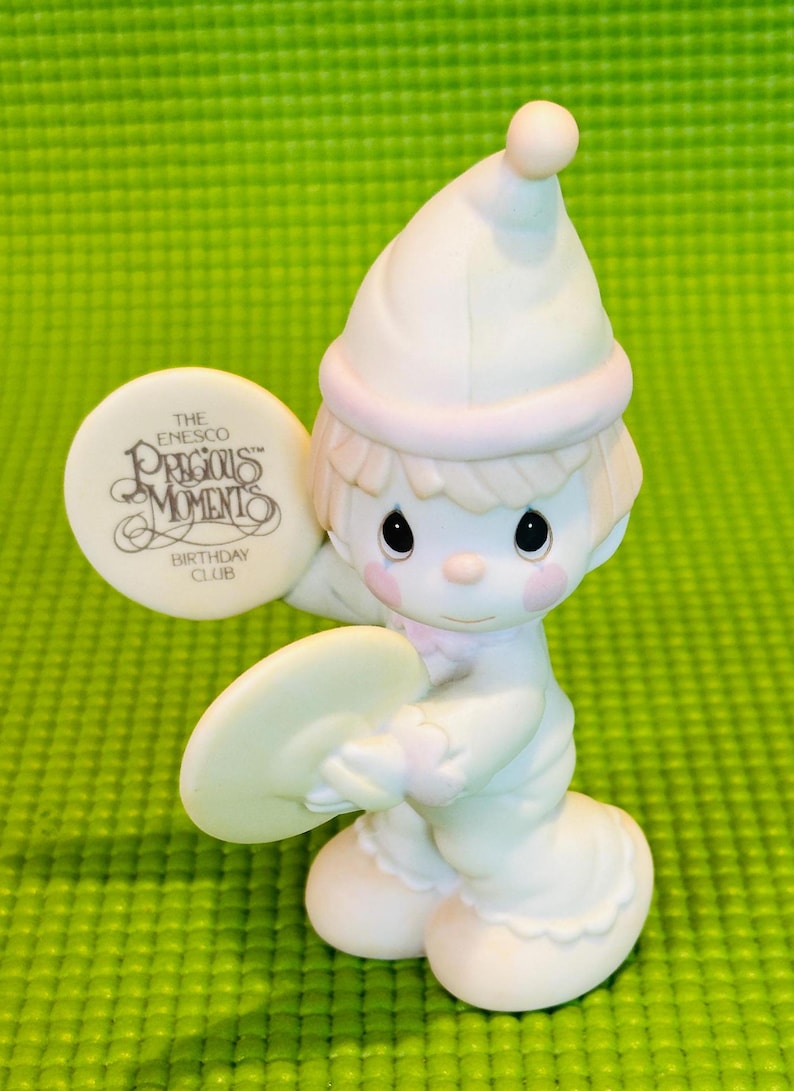May include: A white porcelain figurine of a boy wearing a white and pink hat, holding a white cymbal. The figurine is part of the Precious Moments Birthday Club.
