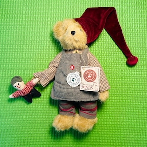 May include: A plush teddy bear wearing a burgundy velvet hat with a long, pointed end. The bear is dressed in a gray overall and plaid shirt, holding a small doll. The bear has a tag that says "Get Yoo Wine Boyds".