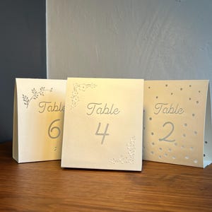 May include: Three ivory table number cards. Each card displays the word "Table" followed by a number: 6, 4, and 2. The cards have decorative floral and dot patterns. The cards are standing on a wooden surface.