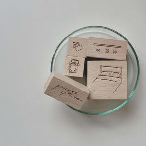 May include: A collection of five light-colored wooden stamps featuring black ink illustrations of a bed, an alarm clock, media playback controls, wireless earbuds, and the handwritten text passage of time, all resting inside a clear glass dish.