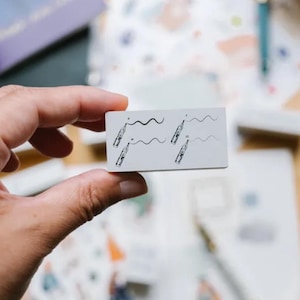 May include: A small, rectangular, white stamp with black line art of pens and wavy lines. The stamp is held in a hand, with other stationery items and stamps visible in the background. The stamp is likely used for crafting or journaling.
