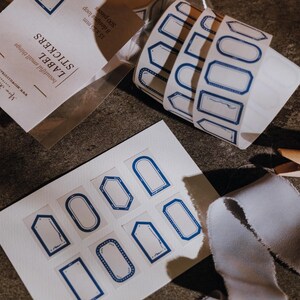 May include: A roll and a sheet of adhesive labels featuring various blue-outlined geometric shapes and frames. The packaging text reads LABEL STICKERS, 15 x 25 mm, 8 designs loop, 500 pieces. The labels are white with blue ink borders.