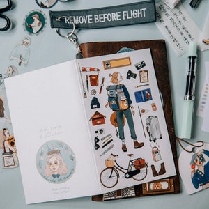 May include: An open notebook with travel-themed stickers, including a backpacker illustration, a bicycle, and luggage. The notebook is surrounded by stickers, a pen, and a keychain that reads "REMOVE BEFORE FLIGHT."
