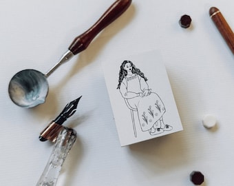 May include: A collection of stationery items is arranged on a white surface. A wooden-handled wax melting spoon and pen are placed near a card with a seated woman illustration. Wax seals and a wooden pen are also visible.