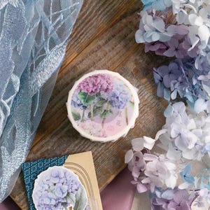 May include: A roll of washi tape with a floral design featuring purple and pink hydrangeas. The tape is on a wooden surface with a blue lace fabric and dried hydrangeas in the background. The tape has the text "紫陽花" in Japanese characters, which translates to "hydrangea".