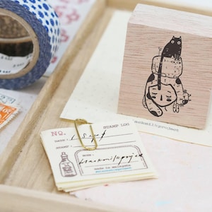 May include: A wooden stamp featuring a black and white illustration of three cats stacked on top of each other. The top cat is sitting upright, while the two cats below are curled up. The stamp is displayed on a wooden surface.
