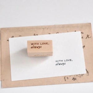 May include: A rectangular wooden rubber stamp featuring the engraved text "WITH LOVE, always" in a serif font. The stamp is positioned on a white card that displays the same text in black ink, resting atop a vintage-style tan envelope.