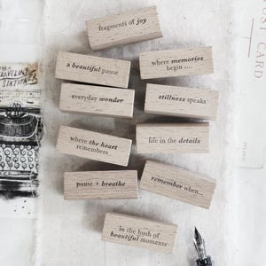 May include: Wooden blocks with phrases such as "fragments of joy" and "a beautiful pause" are displayed on a textured white cloth. The image includes a clip, stationery, and a pen, creating a vintage aesthetic.