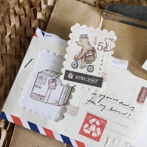 May include: A vintage-style postcard with a handwritten message, "a special drawing for my friend!"  The postcard features three decorative stamps with illustrations of a bear riding a bicycle, a stack of boxes, and a bear holding a package. The stamps are labeled "Kuma Post" and have a 5c denomination.