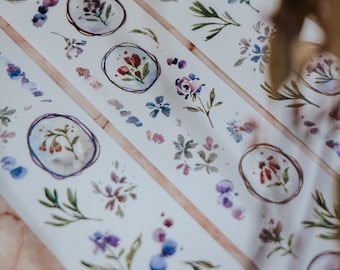 May include: Two rolls of washi tape with floral designs. The tape is white with watercolor illustrations of flowers, leaves, and branches. The tape is labeled "pinot noir" and "Mews Melbourne Australia".