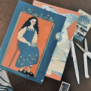 Può includere: Una collezione di articoli di cancelleria su una superficie di legno. Include una cartella blu con un'illustrazione di donna, una cartella fantasia, una penna, forbici, un francobollo e un biglietto dell'autobus. La donna indossa un vestito blu.